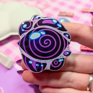 May include: A purple and black swirl pin with a white outline. The swirl has a black center and a white glow around the edges.