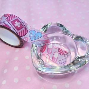 May include: A roll of pink and white washi tape with a medical theme. The tape features illustrations of bandages, pills, hearts, and a heartbeat monitor. The tape is partially unwound and is sitting on a pink surface with white polka dots.