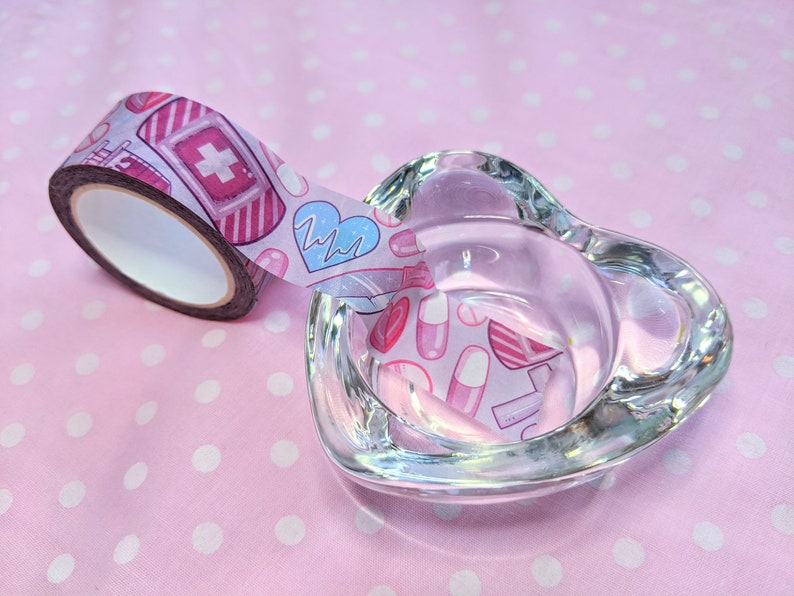 May include: A roll of pink and white washi tape with a medical theme. The tape features illustrations of bandages, pills, hearts, and a heartbeat monitor. The tape is partially unwound and is sitting on a pink surface with white polka dots.
