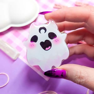 May include: A white ghost with a crescent moon on its head and a big smile. The ghost has pink cheeks and black eyes. The ghost is made of clear acrylic and is a cute and whimsical design.