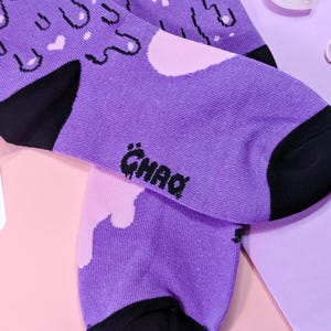 May include: A pair of purple socks with a black and white graphic print of dripping paint and clouds. The socks have a black heel and toe and the word "GHAQ" is printed on the bottom of one sock.