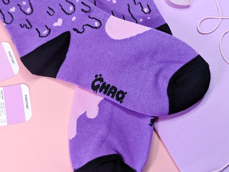 May include: A pair of purple socks with a black and white graphic print of dripping paint and clouds. The socks have a black heel and toe and the word "GHAQ" is printed on the bottom of one sock.