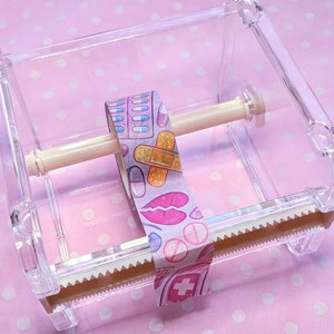 May include: A clear plastic tape dispenser with a roll of patterned washi tape. The tape has a pink background with colorful illustrations of pills, bandages, and lips.