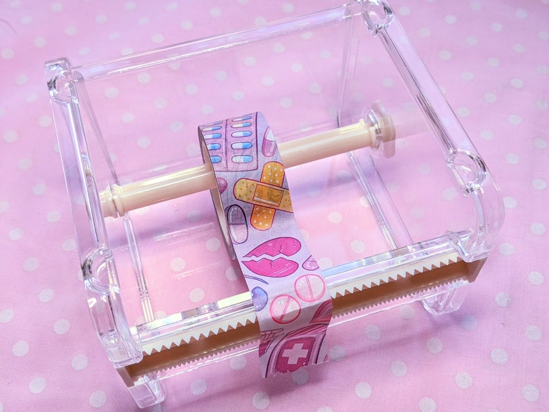 May include: A clear plastic tape dispenser with a roll of patterned washi tape. The tape has a pink background with colorful illustrations of pills, bandages, and lips.