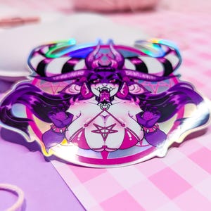 May include: A holographic sticker featuring a purple-haired demon with horns, wearing a pink bikini and a pentagram necklace. The demon is surrounded by a pink and white circle with a pentagram design.