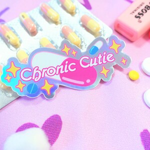 May include: A holographic sticker with a pink pill shape and the text "Chronic Cutie" in white letters. The sticker is surrounded by yellow stars. The sticker is on a pink surface with a blister pack of yellow and orange pills and white pills.