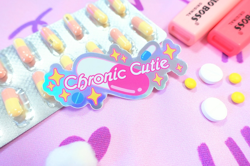 May include: A holographic sticker with a pink pill shape and the text "Chronic Cutie" in white letters. The sticker is surrounded by yellow stars. The sticker is on a pink surface with a blister pack of yellow and orange pills and white pills.