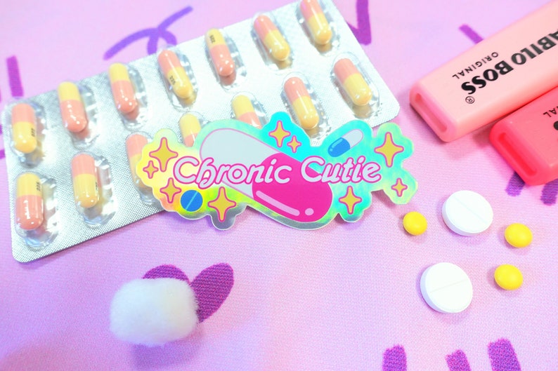 May include: A pink and blue iridescent sticker with the text "Chronic Cutie" and a stylized pill design. The sticker is on a pink surface with a blister pack of pink and yellow pills, two pink highlighters, and white and yellow pills.