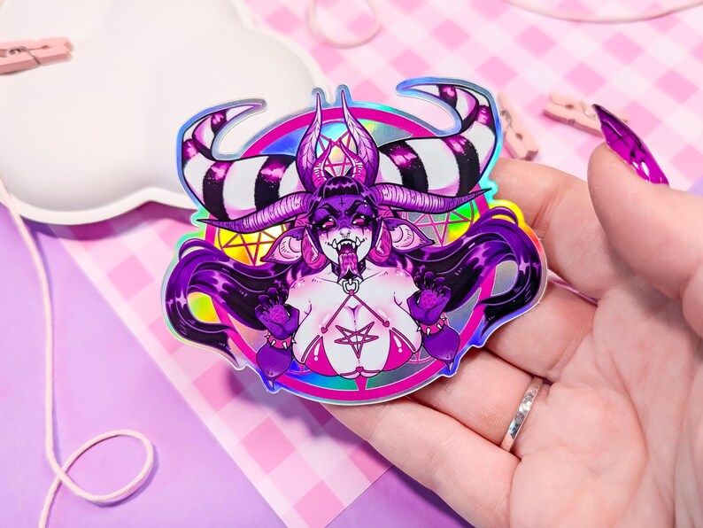 May include: A holographic sticker featuring a purple and black demon with horns, a pentagram, and a pink and white bikini. The demon is surrounded by a rainbow halo.