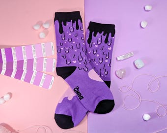 Può includere: Un paio di calzini viola con un design nero che cola e cuori rosa. I calzini hanno la parola "CHAD" scritta su uno di essi.