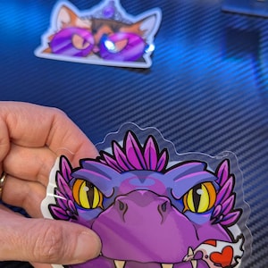 May include: A purple and pink sticker featuring a cartoon dragon with yellow eyes and a heart-shaped object in its mouth. The sticker has a clear border. Another sticker with two cartoon cat faces is visible in the background.