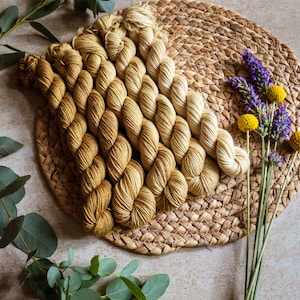 Könnte beinhalten: Ein Set mit fünf Knäuel Garn in einem Farbverlauf von Gelb und Creme. Das Garn ist auf einer geflochtenen Strohmatte mit Zweigen von Eukalyptus und Lavendelblüten angeordnet.