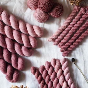 May include: A collection of dusty rose and mauve yarn in various forms, including several twisted hanks, small balls of yarn, and mini skeins. The yarn displays a gradient of pink and muted red tones, arranged alongside dried flower heads.