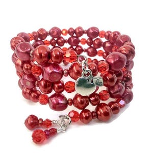 May include: A multi-strand wrap bracelet featuring deep red, irregularly shaped pearls interspersed with small, faceted red crystal beads. A silver-toned metal charm shaped like an apple with a single leaf hangs from the center of the bracelet.