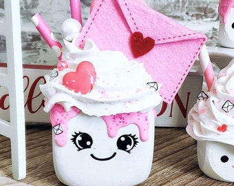 May include: A white ceramic mug with a pink and white whipped cream topping, a pink envelope with a red heart, and two pink and white striped straws. The mug has a pink glaze and a cute cartoon face with black eyes and a smile.