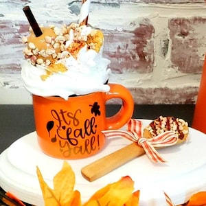 May include: An orange mug with the text "It's Fall Y'all!" is filled with a whipped cream and caramel dessert, topped with nuts and a straw. Two wooden spoons with chocolate and nut toppings are tied with red and white ribbon. Autumn leaves and an orange vase complete the fall theme.