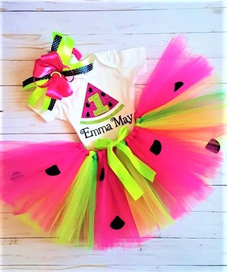 May include: A watermelon-themed birthday outfit for a baby girl. The outfit includes a white onesie with a watermelon slice design and the text "Emma May" and a multi-coloured tulle tutu with black watermelon seeds.