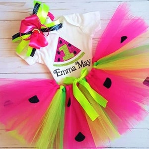 May include: A watermelon-themed first birthday outfit for a baby girl. The outfit includes a white bodysuit with a watermelon slice graphic and the name "Emma May" embroidered on it, a pink and green tulle tutu with black watermelon seeds, and a pink and green bow.