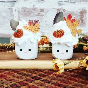 May include: Two white, smiling marshmallow-shaped decorations with whipped cream, orange pumpkins, and fall leaf accents. The decorations sit on a wooden surface with a patterned fabric background, evoking a festive autumn theme.