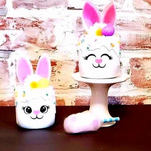 May include: Two white, bunny-themed decorative objects. One sits on a white pedestal, the other on a dark surface. Both have pink ears, a smiling face, and colorful sprinkles. A cotton candy-like object rests near the pedestal.
