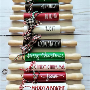 May include: A collection of decorative rolling pins with wooden handles and painted barrels. Each pin features a different holiday-themed phrase in white lettering, such as "Rudolph," "Hot Cocoa," and "Merry Christmas." The pins are adorned with festive ribbons.
