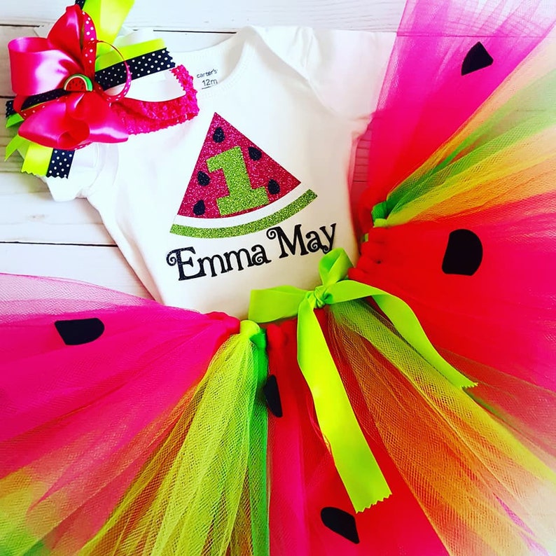 May include: Watermelon-themed first birthday outfit for a baby girl. The outfit includes a white bodysuit with a watermelon slice graphic and the text "Emma May", a pink and green tulle tutu with black watermelon seeds, and a pink headband with a bow and a watermelon slice.