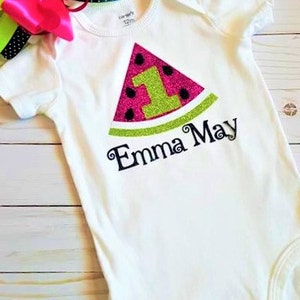 May include: White onesie with a pink and green watermelon design. The watermelon has a green glitter number one and the text "Emma May" is embroidered below.