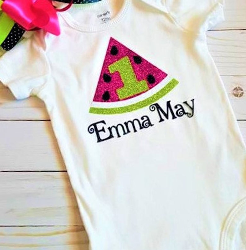 May include: White onesie with a pink and green watermelon design. The watermelon has a green glitter number one and the text "Emma May" is embroidered below.