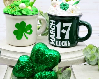 Peut inclure: Deux tasses vertes avec des trèfles blancs. Une tasse a un trèfle blanc et l'autre a le texte "March 17 Lucky". Les deux tasses ont de la crème fouettée blanche avec des pépites vertes et une pièce d'or sur le dessus.