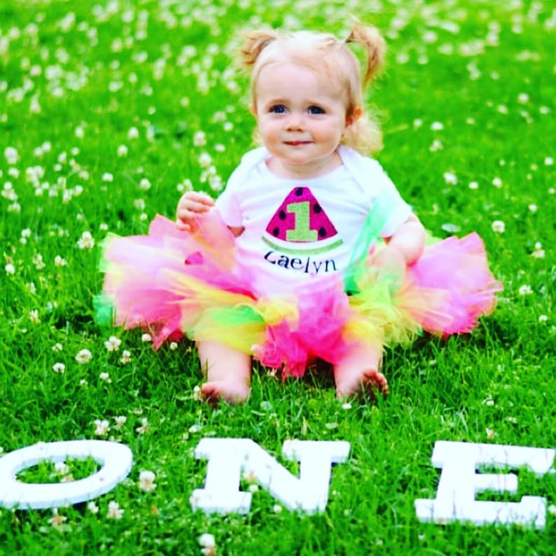 May include: A baby girl wearing a white onesie with a watermelon design and the number one on it. The onesie says "Caelyn" on the bottom. The baby is wearing a pink, yellow, and green tutu and sitting on green grass with white letters spelling out "ONE" in front of her.
