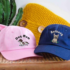 May include: Two baseball caps. One is pink with cat ears, the text "Dog Mum", and a dachshund illustration. The other is navy blue with the name "Anthony" and a French bulldog illustration. A yellow knitted item is in the background.