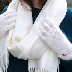 Puede incluir: Una bufanda de cachemira blanca con flecos y un par de guantes de cachemira blancos con las iniciales "KL" bordadas en rosa.