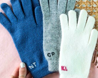 Puede incluir: Tres pares de guantes de punto en azul, gris y blanco con iniciales bordadas. Los guantes azules tienen las iniciales "NT" bordadas en hilo blanco. Los guantes grises tienen las iniciales "CP" bordadas en hilo negro. Los guantes blancos tienen las iniciales "KL" bordadas en hilo rosa.