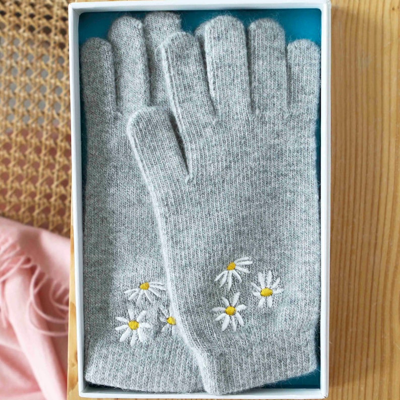 K&ouml;nnte beinhalten: Ein Paar graue Strickhandschuhe mit gestickten wei&szlig;en G&auml;nsebl&uuml;mchen. Die Handschuhe befinden sich in einer wei&szlig;en Schachtel mit blauer Auskleidung.