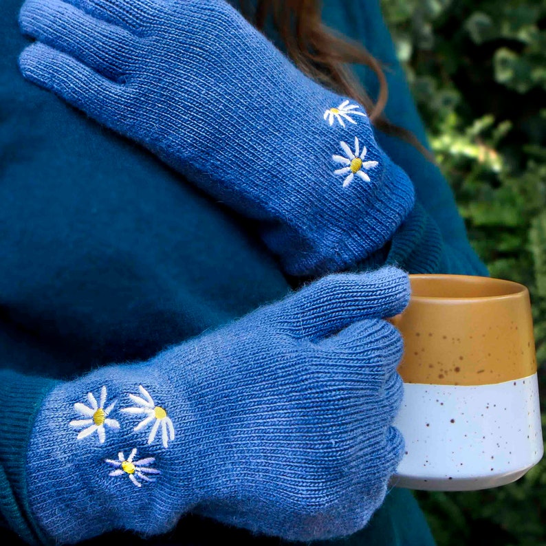 K&ouml;nnte beinhalten: Ein Paar blaue Strickhandschuhe mit wei&szlig;en gestickten G&auml;nsebl&uuml;mchen. Die Handschuhe werden von einer Person in einem t&uuml;rkisfarbenen Pullover gehalten.