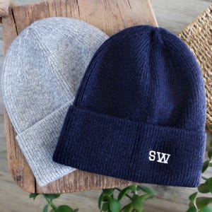 Könnte beinhalten: Zwei Strickmützen. Eine ist hellgrau, die andere marineblau mit den weißen Initialen "SW", die auf den umgeschlagenen Rand gestickt sind. Die Mützen werden auf einer Holzoberfläche präsentiert.