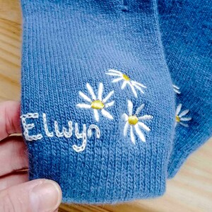 K&ouml;nnte beinhalten: Blaue Socken mit wei&szlig;en gestickten G&auml;nsebl&uuml;mchen und dem Namen "Elwyn" auf der Ferse.