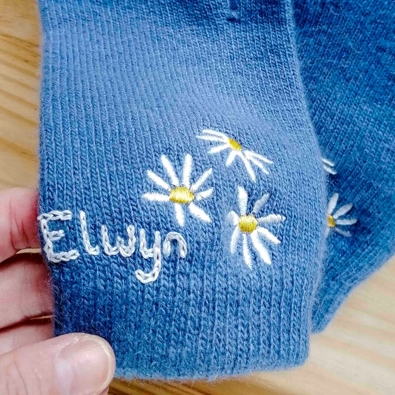 K&ouml;nnte beinhalten: Blaue Socken mit wei&szlig;en gestickten G&auml;nsebl&uuml;mchen und dem Namen "Elwyn" auf der Ferse.
