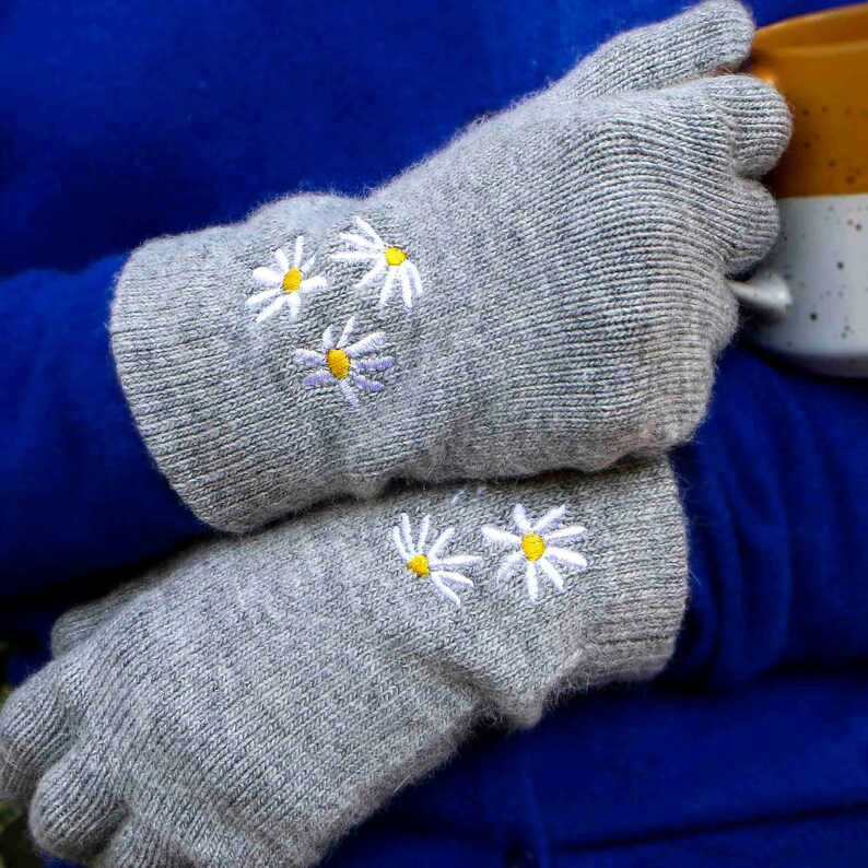 K&ouml;nnte beinhalten: Ein Paar graue Strickhandschuhe mit wei&szlig;er G&auml;nsebl&uuml;mchen-Stickerei. Die G&auml;nsebl&uuml;mchen haben gelbe Zentren.
