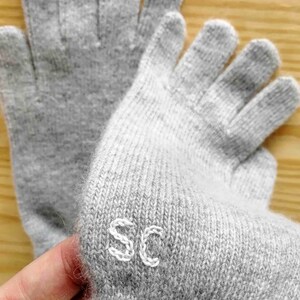 Puede incluir: Un par de guantes sin dedos grises con costuras blancas. Los guantes tienen las letras "SC" bordadas en la palma.