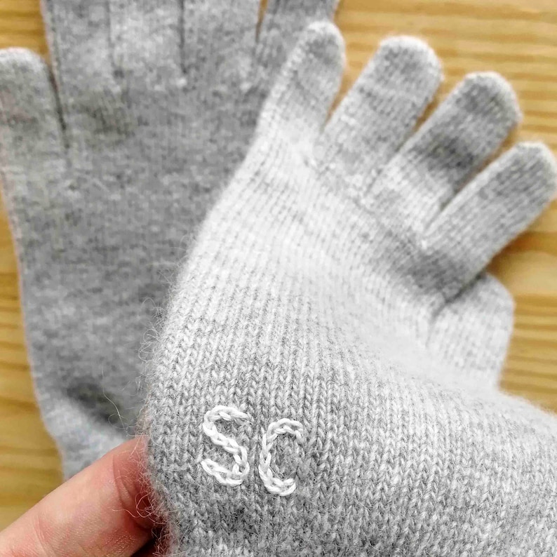 Puede incluir: Un par de guantes sin dedos grises con costuras blancas. Los guantes tienen las letras "SC" bordadas en la palma.