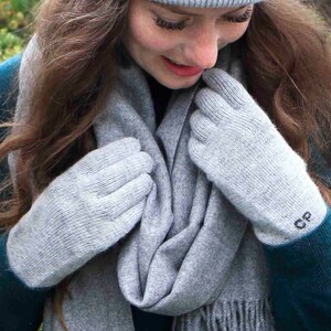 Puede incluir: Un par de guantes de punto grises con un peque&ntilde;o monograma "CP" en un guante. Los guantes est&aacute;n siendo sostenidos por una persona que lleva una bufanda gris y un gorro gris.