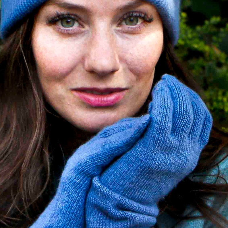 Puede incluir: Una mujer con un gorro azul de punto y guantes azules de punto a juego. Los guantes se sostienen juntos frente a su cara.