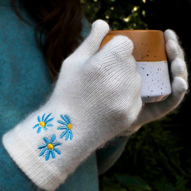 K&ouml;nnte beinhalten: Wei&szlig;e Strickhandschuhe mit blauen und gelben gestickten Blumen. Die Handschuhe werden von einer Person gehalten, die eine Tasse h&auml;lt.