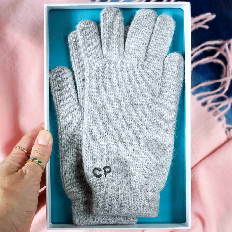 Puede incluir: Un par de guantes de punto grises con las iniciales "CP" bordadas en la parte posterior de un guante. Los guantes est&aacute;n en una caja blanca con fondo azul.