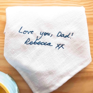 May include: White handkerchief with blue embroidered text that reads "Love you, Dad! Rebecca xx". The handkerchief is folded and lies on a wooden surface. The embroidery is in a handwritten style.