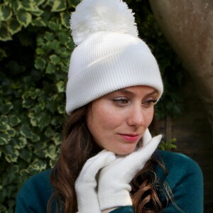K&ouml;nnte beinhalten: Eine wei&szlig;e Strickm&uuml;tze mit einem flauschigen wei&szlig;en Bommel oben. Die M&uuml;tze wird von einer Person mit langen braunen Haaren getragen, die wei&szlig;e Handschuhe tr&auml;gt.