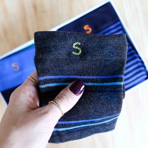 May include: A pair of dark gray socks with blue stripes and an embroidered green "S" on the top of the sock. The socks are folded and held in a hand.