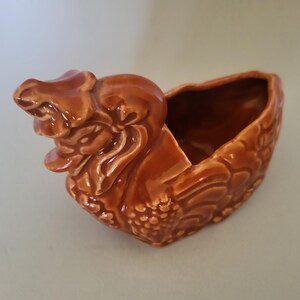 May include: A brown ceramic planter in the shape of a mythical creature. The planter has a detailed design with a textured surface and a glossy finish. The open container is approximately 6 inches long.