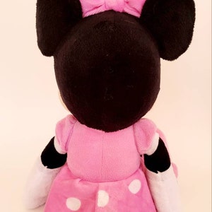 Op de afbeelding: Een pluche Minnie Mouse pop, van achteren gezien, met een zwarte kop, grote oren en een roze strik. De pop draagt een roze jurk met witte stippen en heeft zwart-witte accenten op de armen.